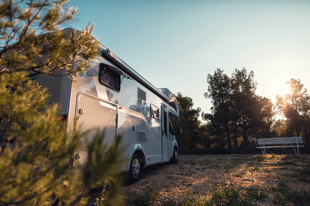 A white RV is parked on a grassy area near trees, with the sun setting in the background.